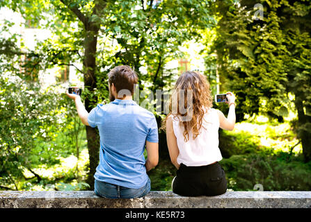 Bella coppia Giovane con smart phone seduto sul muro di cemento, tenendo selfies. soleggiata giornata di primavera. vista posteriore. Foto Stock