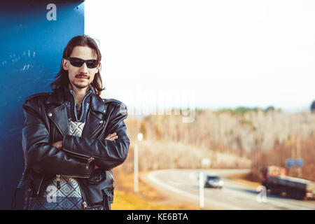 Biker maschio in una giacca di pelle e bandana mette su occhiali da sole e guardando la telecamera. motociclista in giacca di pelle mette in occhiali da sole. Foto Stock