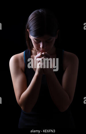 Fedeli athletic donna orante, con mani giunte nel culto a Dio, a testa in giù e gli occhi chiusi in fervore religioso, sulla chiave di basso sfondo nero. Foto Stock