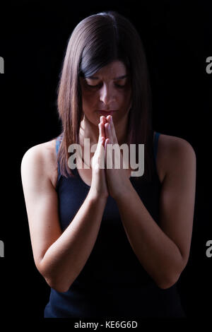 Fedeli athletic donna orante, con mani giunte nel culto a Dio, a testa in giù e gli occhi chiusi in fervore religioso, su sfondo nero. concetto per Foto Stock