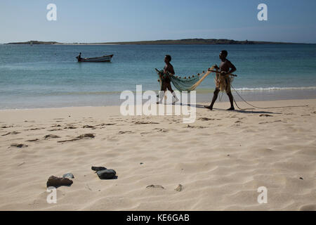 Due pescatori a camminare su una spiaggia di Capo Verde Isola azienda una rete da pesca con una piccola barca da pesca in background Foto Stock