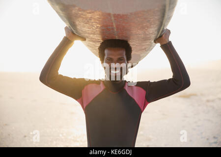 Ritratto sorridente, sicuro surfista maschile che porta surf in alto sulla soleggiata spiaggia estiva Foto Stock