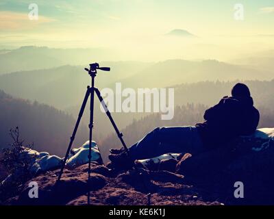 Natura professionale fotografo fare il lavoro su terreni innevati cliff. l uomo si prende le foto con la fotocamera dello specchietto sul picco di roccia. sognante paesaggio nebbiosi, molla pi arancione Foto Stock