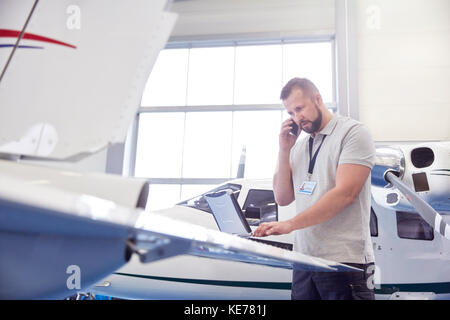 Meccanico di aeroplano maschio che parla sul telefono delle cellule e che lavora a. computer portatile in hangar Foto Stock