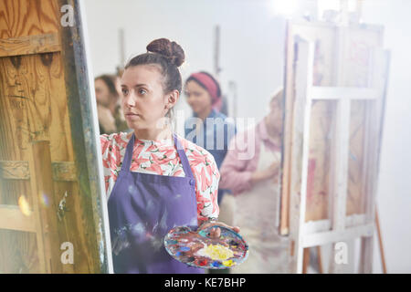 Artista femminile focalizzato con pittura di tavolozza a easel in arte studio di classe Foto Stock