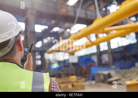 Lavoratore maschile che utilizza walkie-talkie per guidare l'attrezzatura di abbassamento della gru idraulica in fabbrica Foto Stock