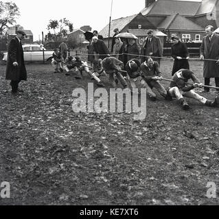 Anni sessanta, storico rimorchiatore guerra iof contest, su un campo fangoso a un club sportivo locale, il maschio coach monitora il suo team come essi tutti tirare insieme sulla fune, Inghilterra, Regno Unito. Foto Stock