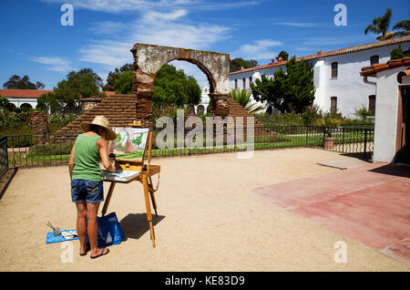 Donna della pittura la scena su un cavalletto in un cortile presso la missione di San Luis Rey, vicino Oceanside CA in tarda estate; California, Stati Uniti d'America Foto Stock