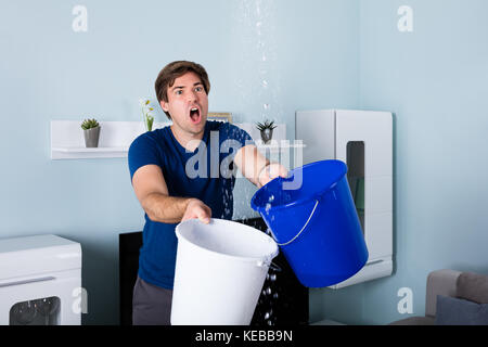 Giovane Maschio uomo preoccupato tenendo la benna mentre le goccioline di acqua che perde dal soffitto Foto Stock