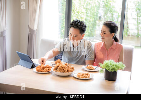 Giovani coppie asiatiche mangiare pollo fritto insieme nel salotto di casa contemporanea per lo stile di vita moderno concetto Foto Stock