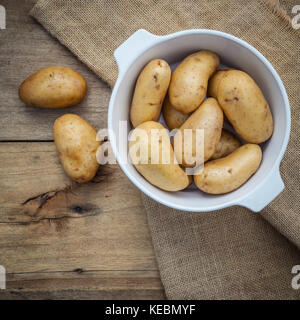 Composizione di freschi Patate organico in ceramica bianca ciotola sulla canapa sake e tavola in legno rustico. Preparazione per la cottura. Foto Stock