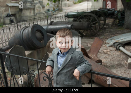 Un caucasian little boy in giacca e camicia in un museo navale con le pistole e delle eliche sullo sfondo Foto Stock