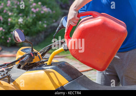 Uomo caucasico nello sport occhiali protettivi detiene il barattolo rosso e versa la benzina in atv quadbike del serbatoio. processo di rifornimento di carburante. le operazioni di manutenzione e riparazione. Foto Stock