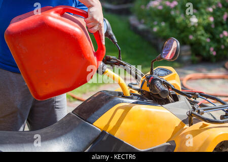 Uomo caucasico nello sport occhiali protettivi detiene il barattolo rosso e versa la benzina in atv quadbike del serbatoio. processo di rifornimento di carburante. le operazioni di manutenzione e riparazione. Foto Stock