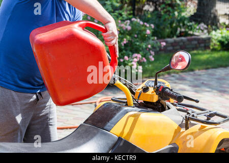 Uomo caucasico nello sport occhiali protettivi detiene il barattolo rosso e versa la benzina in atv quadbike del serbatoio. processo di rifornimento di carburante. le operazioni di manutenzione e riparazione. Foto Stock