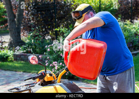 Uomo caucasico nello sport occhiali protettivi detiene il barattolo rosso e versa la benzina in atv quadbike del serbatoio. processo di rifornimento di carburante. le operazioni di manutenzione e riparazione. Foto Stock