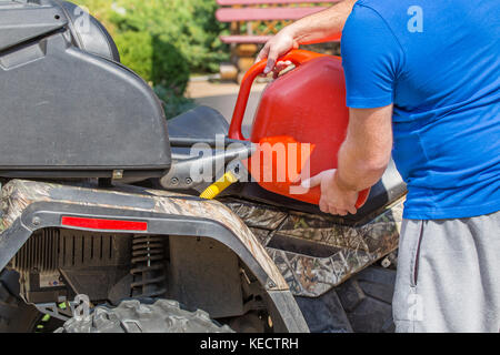 Uomo caucasico nello sport occhiali protettivi detiene il barattolo rosso e versa la benzina in atv quadbike del serbatoio. processo di rifornimento di carburante. le operazioni di manutenzione e riparazione. Foto Stock