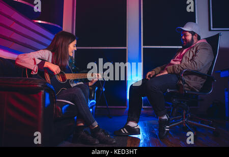 La donna a suonare la chitarra e cantare con uomo seduto su una sedia in ascolto. Musicisti di comporre una canzone sulla chitarra in studio di registrazione. Foto Stock