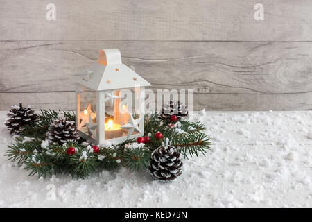 Bianco Natale lanterna con candela accesa su abete ramoscelli decorate con agrifoglio bacche, pigne e fiocchi di neve. Foto Stock