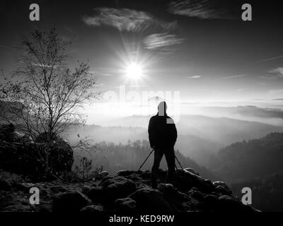 Mattina di sole con la prima neve. fotografo preparare la fotocamera sul cavalletto. rocce innevate, nella valle di foglie colorate foresta. Vista su misty e fogg Foto Stock