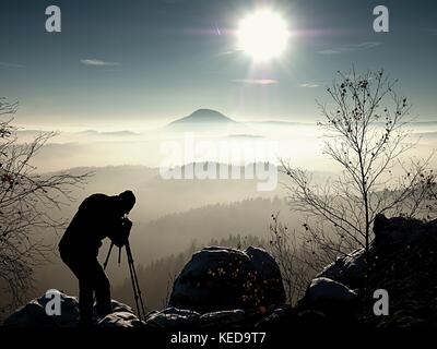 Mattina di sole con la prima neve. fotografo preparare la fotocamera sul cavalletto. rocce innevate, nella valle di foglie colorate foresta. Vista su misty e fogg Foto Stock