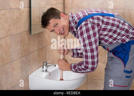 Sorridente Giovane Maschio idraulico utilizzando lo stantuffo in ostruito il lavandino in bagno Foto Stock