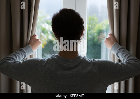 Vista posteriore della donna che guarda attraverso la finestra a casa Foto Stock