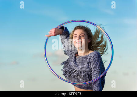 Ragazza sorride come lei esegue la cerchiatura, catturato a metà in aria, guardando attraverso hoop contro un cielo blu con alcune piccole nuvole. Foto Stock