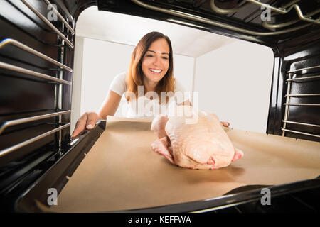 Giovane donna felice indossando i guanti ponendo il vassoio di pollo al forno Foto Stock