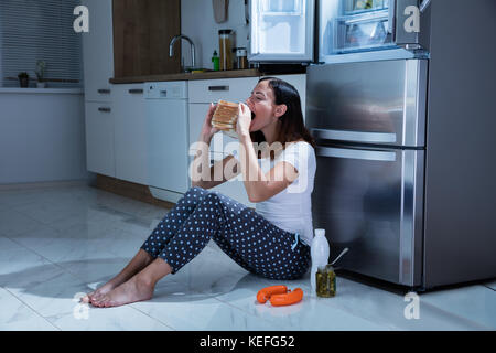 Giovane donna mangiare panino con un vasetto di sottaceti mentre è seduto sul pavimento in cucina Foto Stock