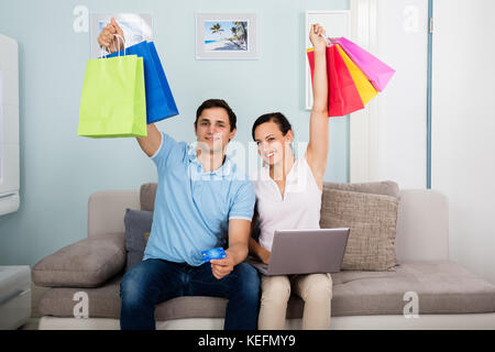 Sorridente giovane seduto sul divano con il computer portatile azienda multi-colore di borse per lo shopping Foto Stock