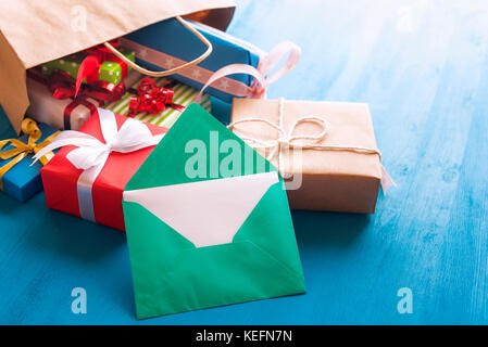 Busta verde con vuoto di carta bianca appoggiata contro un mazzetto di colorato presenta, che escono da un marrone shopping bag, su un blu sullo sfondo di legno Foto Stock