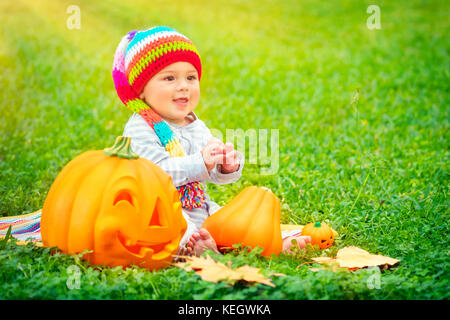 Carino piccolo bambino seduto sul prato verde campo indossando divertente colorato hat e giocando con le zucche con sorridente volti scolpiti, happy Halloween holiday Foto Stock