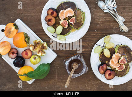 Set colazione. in casa zucchini pancakes con prugna fresca, mandarino, uva, fichi e miele in bianco di piastre in ceramica su legno rustico sfondo, per Foto Stock