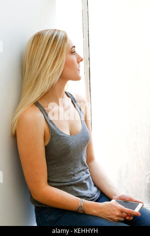 Donna caucasica pensiva seduta sul windowsill che tiene il telefono delle cellule Foto Stock