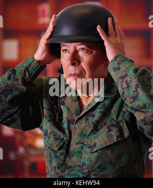 Close up di un grave soldato veterano che indossa un casco e una uniforme militare, con entrambe le mani la regolazione del casco, in uno sfondo sfocato Foto Stock