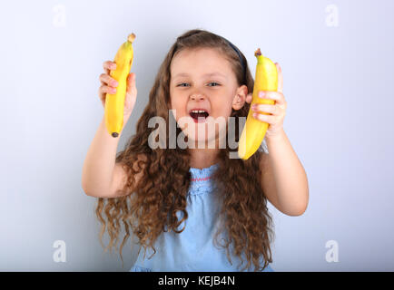 Giocoso ridendo felice capelli lunghi kid girl holding e mostrando il giallo luminoso banane su sfondo blu Foto Stock