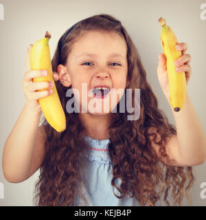 Giocoso ridendo felice capelli lunghi kid girl holding e mostrando il giallo luminoso banane. tonico ritratto vintage Foto Stock