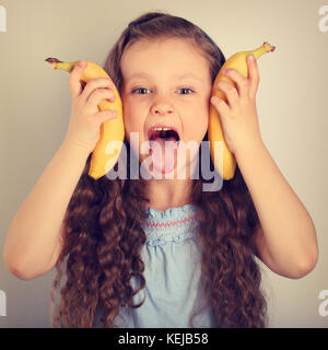 Giocoso divertimento capelli lunghi kid ragazza che mostra la linguetta e azienda brigh giallo banane. tonico ritratto vintage Foto Stock