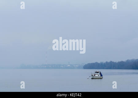 Belgrado sotto la nebbia visto da zemun, con una barca di fronte, sul fiume Danubio e i fumi di un camino industriale nell'immagine di sfondo o Foto Stock