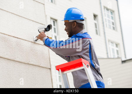 Giovane maschio felice tecnico su scala riparazione telecamera di sorveglianza Foto Stock