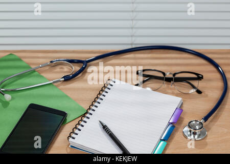Lo stetoscopio e un blocco note la posa su un banco medici con uno smart phone e una tenda a veneziana caduta indietro Foto Stock