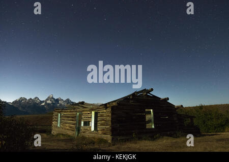 Un vecchio abbandonati cabina homesteader home siede sotto un solitario Starlit Sky con il Grand Teton Mountains del Wyoming in distanza. Foto Stock