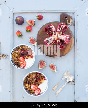Sana colazione set. bocce di oat granola con yogurt, fragole fresche, fichi, melograni ed il miele sulla luce blu sullo sfondo di legno, vista dall'alto Foto Stock