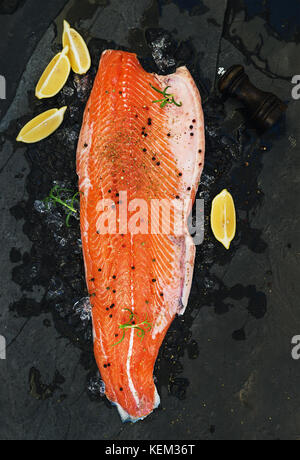 Materie di filetto di salmone con limone e rosmarino sul ghiaccio scheggiata su pietra scura sullo sfondo, vista dall'alto Foto Stock