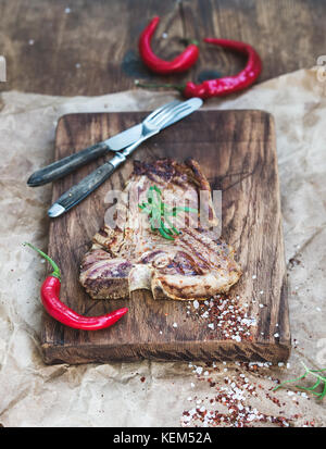 Carne cotta t-bone steak sulla scheda servente con peperoncino rosso, di spezie e di rosmarino fresco su oleoso carta craft, rustico sfondo di legno Foto Stock