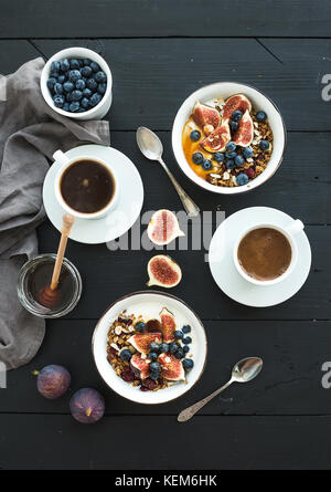Sana colazione set. bocce di oat granola con yogurt, mirtilli freschi e fichi, caffè, miele, nero su uno sfondo di legno Foto Stock