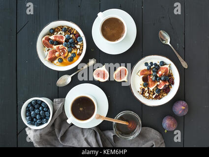 Sana colazione set. bocce di oat granola con yogurt, mirtilli freschi e fichi, caffè, miele, nero su uno sfondo di legno Foto Stock
