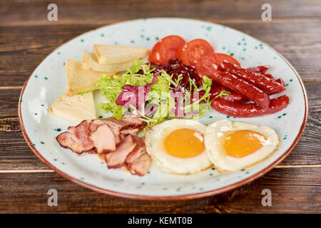 Prima colazione inglese con insalata fresca. prima colazione inglese - uova fritte, pancetta cotta al forno e salsicce, toast, pomodori e insalata verde. appetitoso piatto su pla Foto Stock