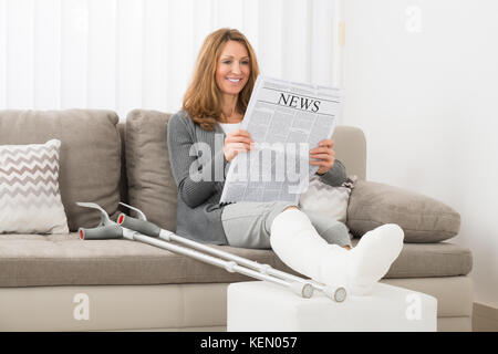 Donna matura con gamba fratturata leggendo il giornale in casa Foto Stock